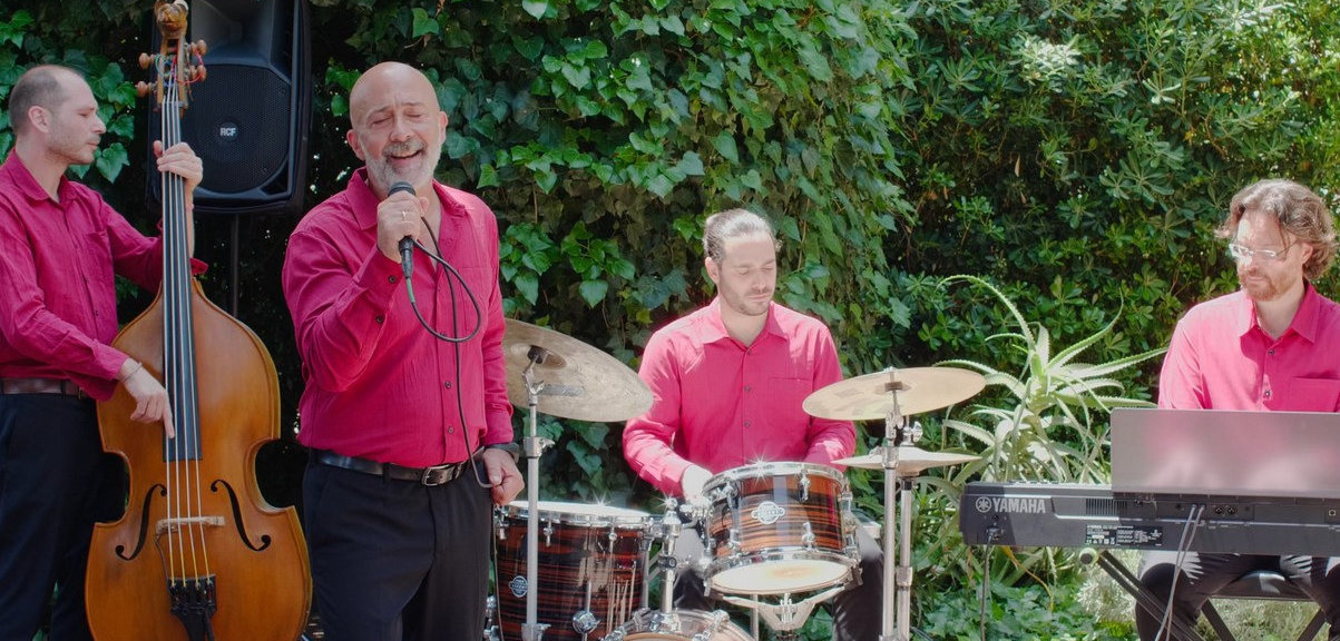 Sarabanda 4tet sul set del video di "L-O-V-E", estate 2025. In camicia rossa bordeaux su sfondo verde (un gazebo di edera e altre piante)
