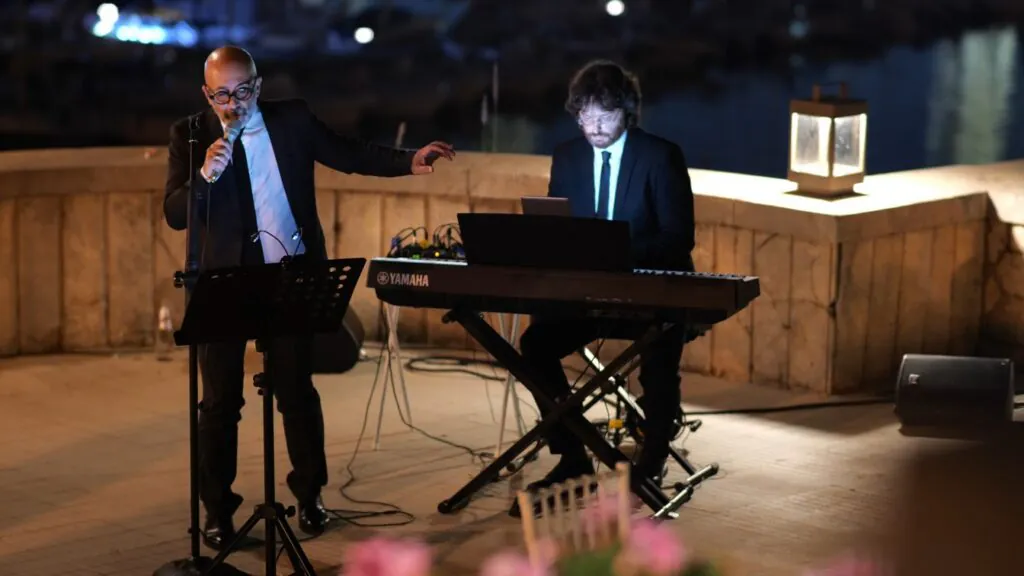 Salvo Samaritano e Roberto Mezzatesta suonano in duo ad un aperitivo di fronte al mare, barche sullo sfondo. Ripresa del totale. Villa Igiea, Palermo.